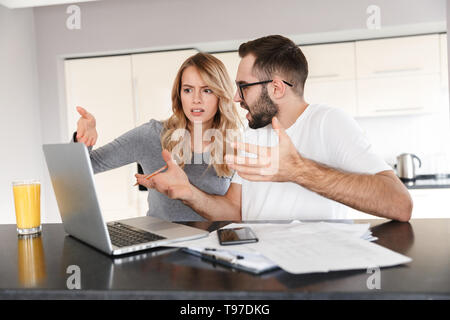 Image of displeased young loving couple sitting at the kitchen using laptop computer. Stock Photo