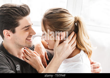 Image of optimistic couple man and woman hugging together while sitting on sofa in bright room at home Stock Photo