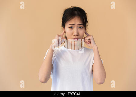 Portrait of upset asian woman wearing basic t-shirt plugging ears with her fingers isolated over beige background in studio Stock Photo