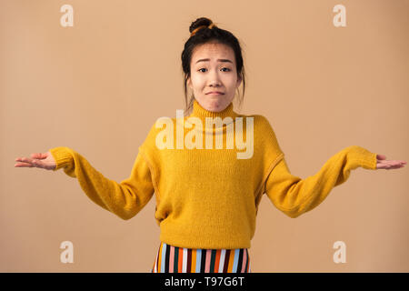Image of innocent japanese woman wearing sweater throwing up hands with puzzlement isolated over beige background in studio Stock Photo