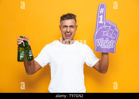 Image of happy man 30s wearing number one fan hand glove with finger raised while drinking beer isolated over yellow background Stock Photo