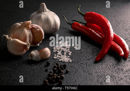 on a black background two heads and cloves of garlic opposite three red chili pepper between them track coarse sea salt and black peppercorns Stock Photo