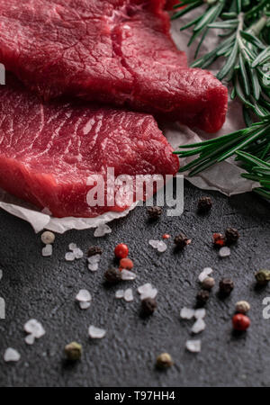 two pieces raw beef steak with fresh rosemary branches lay on parchment paper beside on a dark background scattered large sea salt with red and black  Stock Photo