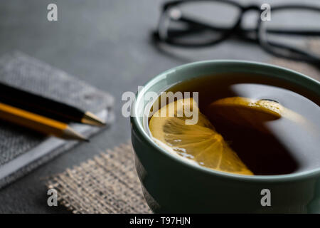Black tea with two pieces of lemon on a light burlap napkin with a small gray notepad, black pen a pencil and glasses Stock Photo