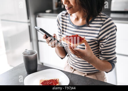 Cropped image of a young african woman having breakfast while sitting at the kitchen, using mobile phone Stock Photo