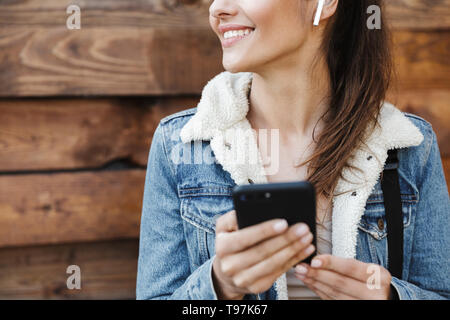 Cropped image of a beautiful young brunette woman wearing jacket, carrying backpack walking outdoors over wooden background, listening to music with e Stock Photo
