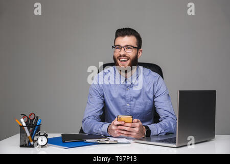 Cheerful bearded elegant man in eyeglasses sitting with smartphone and looking away Stock Photo