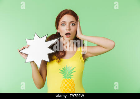 Image of a beautiful shocked scared young redhead girl posing isolated over green wall background holding speech bubble. Stock Photo