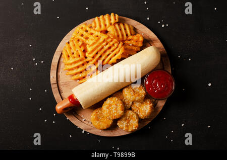 French hot-dog in bun served with ketchup with crispy potato waffles fries and cheese nuggets on a black background Stock Photo