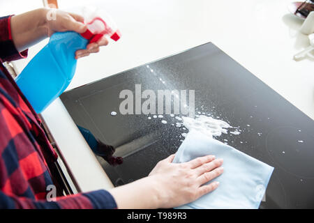 Woman splashes cleaning foam from blue bottle on the induction hob and wipes it using blue cloth. Stock Photo