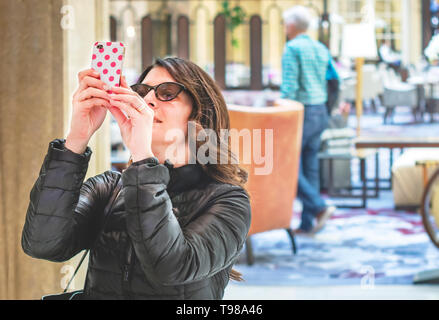 young woman with sunglasses and brown hair taking photographs with her smart phone in an elegant room. Tourism and travel. Pleasant and relaxing activ Stock Photo