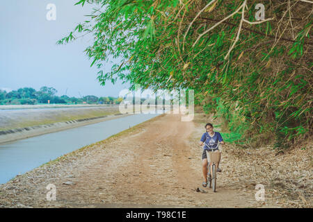 KANCHANABURI THAILAND - APRIL 4 : Unidentified young woman riding a bicycle on gravel road along the irrigation canal at  Mae Klong dam on April 4,201 Stock Photo