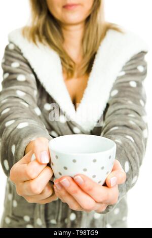 Blonde woman in gray dressing gown with white dots offers white mug with gray dots - coffee or tea - white background Stock Photo
