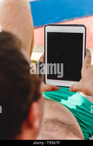 A young man in sunglasses holds a tablet in his hands. Freelancer job concept on the beach and vacation. Stock Photo