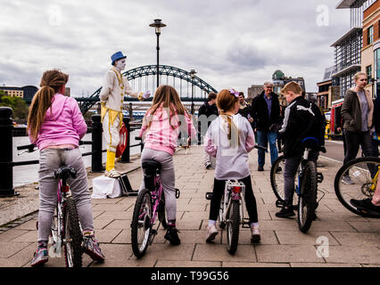 Rear view of group of children riding bikes alongside quay, street entertainer in front of them, Quayside, Newcastle upon Tyne, England, UK Stock Photo