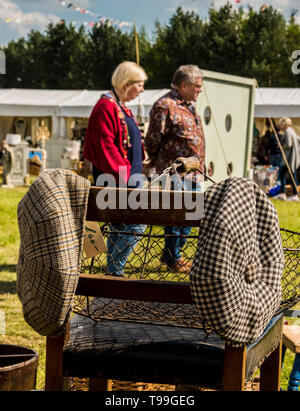Heterosexual couple walking past antique stall, chair with two flat caps hanging on it in foreground, Ripley, North Yorkshire, England, UK Stock Photo