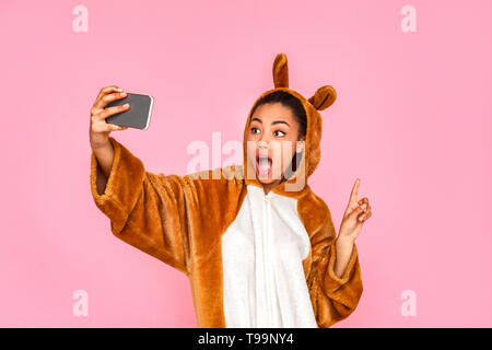 Young woman in bunny kigurumi standing isolated on pink background taking selfie picture on smartphone pointing at space aside mouth opened surprised Stock Photo