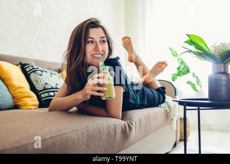 Happy young woman relaxing in living room and drinking smoothie. Healthy diet