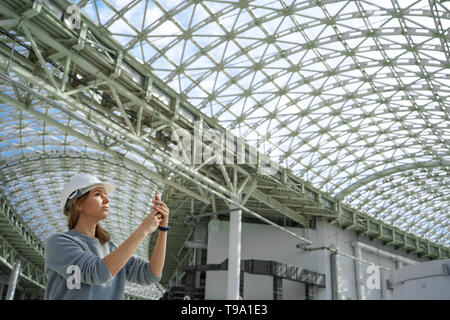 A young girl in a helmet in a large room with a glass roof takes pictures on the phone. A serious expert, a woman inspects the object Stock Photo