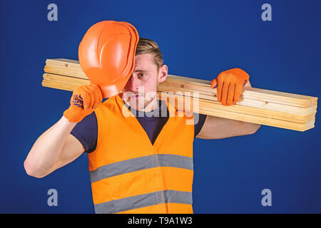 Protective equipment concept. Carpenter, woodworker, labourer, builder carries wooden beam on shoulder. Man in protective gloves holds visor of helmet, corrects hard hat on head, blue background. Stock Photo