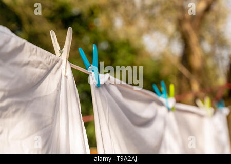 White clothes hung out to dry on a washing line and fastened by the clothes pegs in the bright warm sunny day. Blurred garden at background. Stock Photo