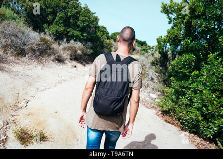 a young caucasian man wearing jeans and a green t-shirt, carrying a backpack, seen from behind, walking by a dirt road Stock Photo