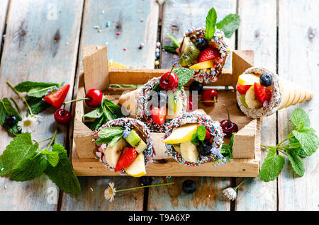 Fruit salad in ice-cream cones, arranged in a small crate on a wooden surface and arranged with mint leaves and lemon balm Stock Photo