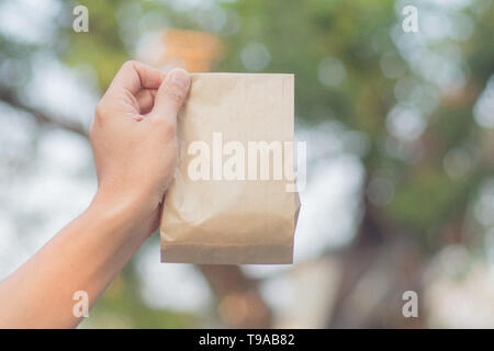 Close up female holds in hand brown clear empty blank craft paper bag for take away on blur bokeh nature background. Packaging bag. Stock Photo