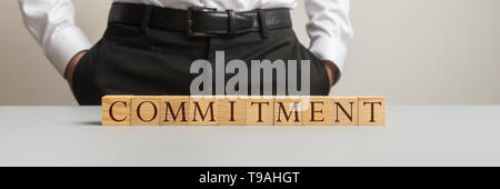Wide view image of businessman standing behind a desk with a Commitment sign assembled of wooden cubes. Stock Photo