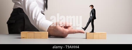 Wide view image of businessman placing his hand between two blocks of wooden cubes for another businessman to walk across in a conceptual image. Stock Photo