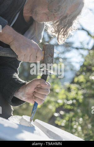Senior artist using mallet and chisel to carve in stone outside on a sunny day. Stock Photo