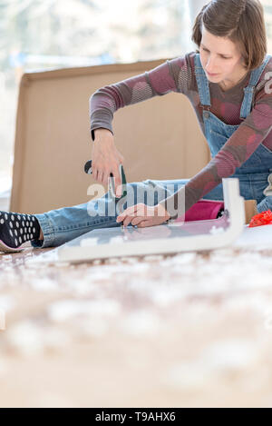 Competent young woman assembling flat pack furniture using a handheld drill as she braces it with her leg for stability. Stock Photo