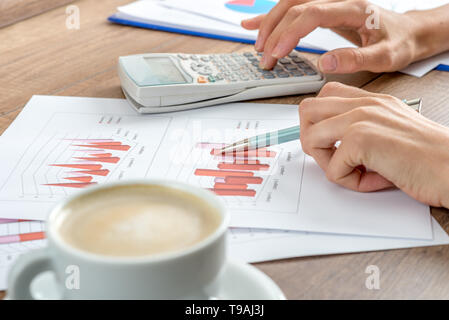 Female accountant working on a presentation or annual report analysing a set of graphs and charts with a manual calculator with a cup of coffee alongs Stock Photo