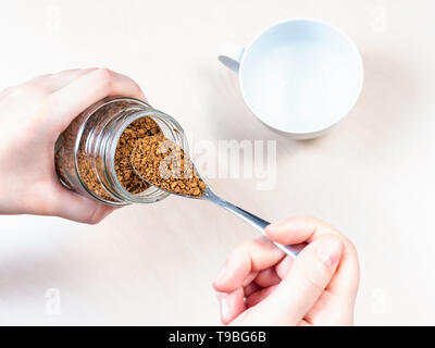 top view of hand spooning up instant coffee from glass jar over cup on light brown table Stock Photo