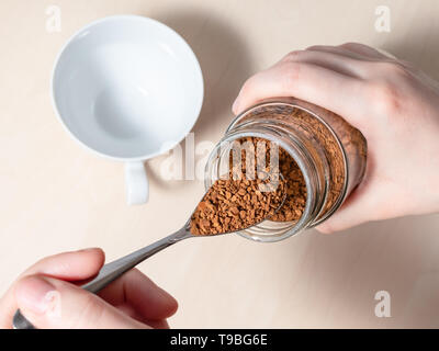female hand spooning up instant coffee from glass jar over cup on light brown table Stock Photo