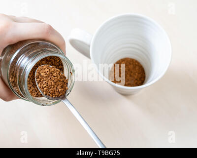 top view spooning up second portion of instant coffee from glass jar over mug on light brown table Stock Photo