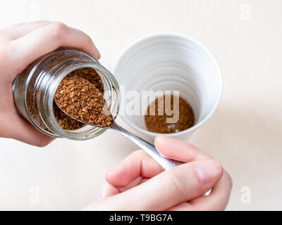 top view of hand spooning up second portion of instant coffee from glass jar over mug on light brown table Stock Photo