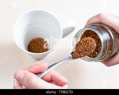 female hand spooning up second portion of instant coffee from glass jar over mug on light brown table Stock Photo