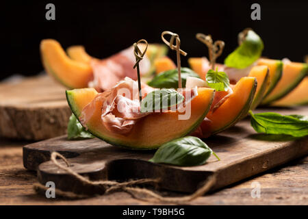 Sliced melon with ham and basil leaves, served on a wood chopping board close up Stock Photo