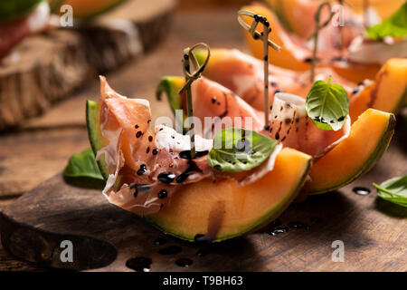 Sliced melon with ham and basil leaves, served on a wood chopping board close up Stock Photo
