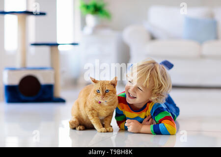 Child playing with cat at home. Kids and pets. Little boy feeding and petting cute ginger color cat. Cats tree and scratcher in living room interior. 
