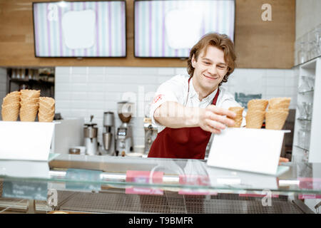 Handsome smiling salesman taking waffle cone while making ice cream for the client in the modern pastry shop Stock Photo