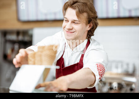 Handsome smiling salesman taking waffle cone while making ice cream for the client in the modern pastry shop Stock Photo