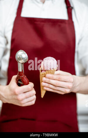 Salesman holding ice cream in the waffle cone and professional scoop on the red apron background, close-up view Stock Photo
