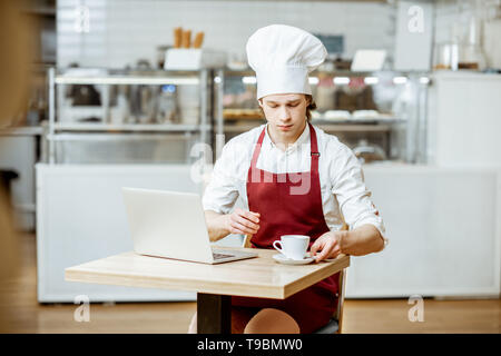 Chef cook in apron and hat working with laptop and drinking coffee in the pastry shop or cafe Stock Photo