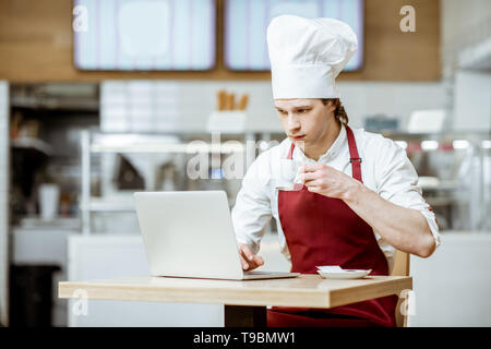 Chef cook in apron and hat working with laptop and drinking coffee in the pastry shop or cafe Stock Photo