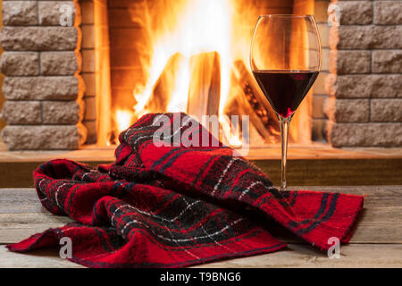 Glass of red wine and warm scarf against cozy fireplace background, in country house, horizontal, hygge , home sweet home. Stock Photo