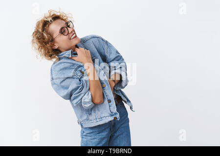 Indoor shot of excited happy and carefree stylish young european woman with shrot curly hairstyle turning right wearing glasses putting denim jacket Stock Photo