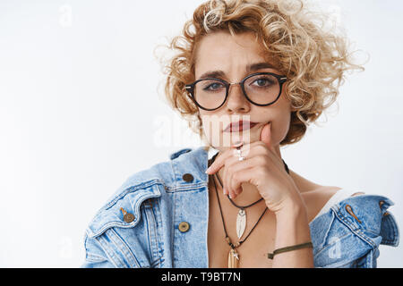 Questioned girl looking doubtful and suspicious. Portrait of uncertain attractive girlfriend in glasses and denim jacket holding hand on chin Stock Photo