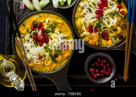laksa with pumpkin and coconut milk, rice noodles, broccoli and pomegranate seeds in portioned black plates on a dark background. healthy vegan dish Stock Photo